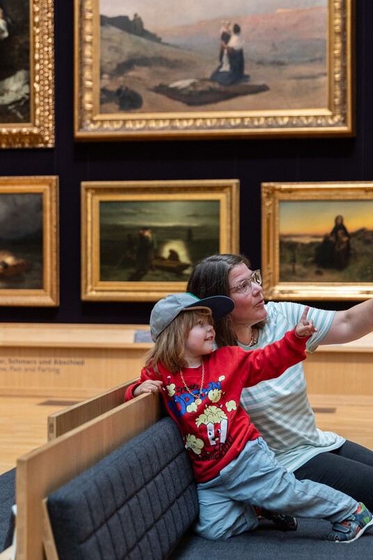 Mutter und Sohn sitzen auf einer Bank im Ausstellungsraum und zeigen auf Gemälde